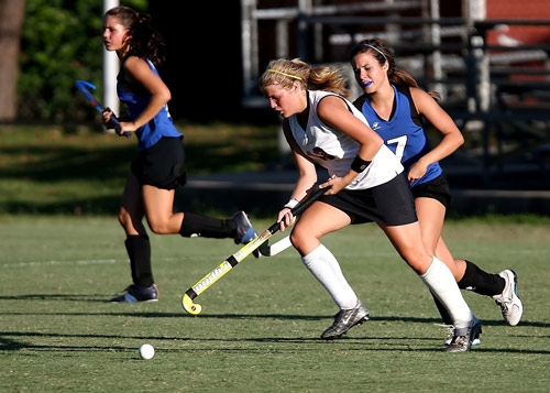 Image of field hockey players playing the sport wearing some of the required field hockey equipment, including hockey stick, ball, mouth guard, shoes, socks, arm protector, and leg guards.